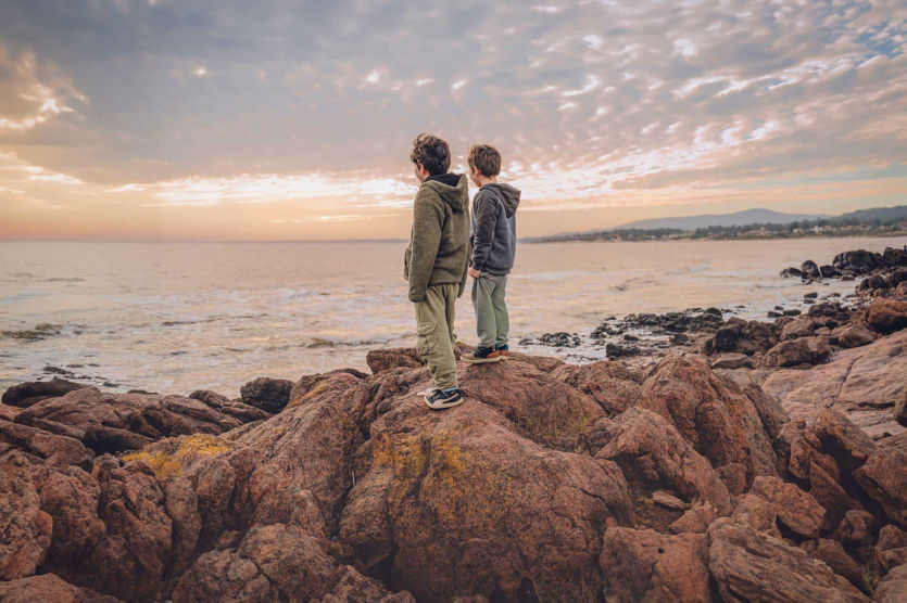 Dos niños de espaldas sobre rocas mirando el mar al atardecer en la costa de Uruguay – fotografía infantil natural por Caro Roig.
