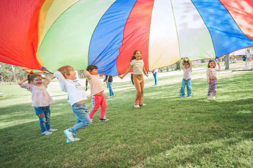 Grupo de niños jugando con lona multicolor en una fiesta de cumpleaños al aire libre.